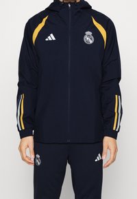 Marineblauw trainingspak met gele en paarse strepen, met Real Madrid- en Adidas-logo's op jas en broek, gedragen door een staand persoon.