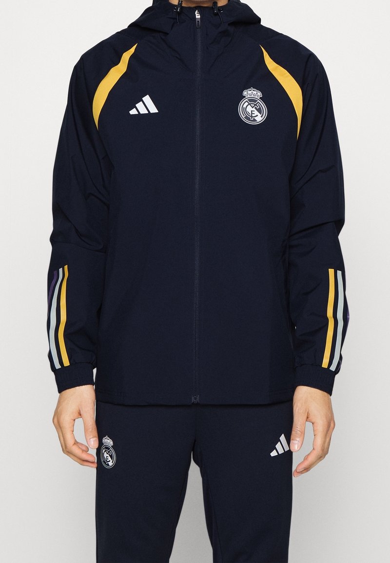 Marineblauw trainingspak met gele en paarse strepen, met Real Madrid- en Adidas-logo's op jas en broek, gedragen door een staand persoon.