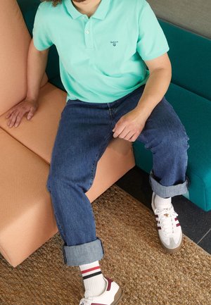 Person trägt türkisfarbenes Poloshirt, hochgekrempelte blaue Jeans, weiße Sneakers mit kastanienbraunen Streifen, sitzt auf einem zweifarbigen Sofa mit orangefarbenen und türkisfarbenen Kissen.