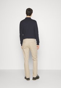 Uomo di fronte a un muro bianco, indossa una maglietta nera a maniche lunghe, pantaloni beige e scarpe nere, in piedi su un pavimento grigio chiaro.