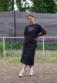 Camiseta negra con "LIONESSES" y el logo de Nike, combinada con una falda de mezclilla larga y zapatillas blancas con acentos rojos. Entorno al aire libre.