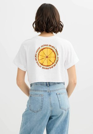 Mujer con cabello corto y castaño lleva una camiseta blanca corta que muestra una rodaja de cítrico naranja y el texto "Fresh Citrus Splash" dentro de un círculo en la parte trasera.