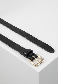 Cinturón de cuero negro con una hebilla de metal dorado, textura suave, múltiples agujeros para ajustar, colocado en una superficie clara.