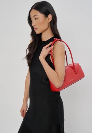 Femme en robe noire sans manches tenant un sac à main rouge Lancaster sur son épaule devant un fond uni.