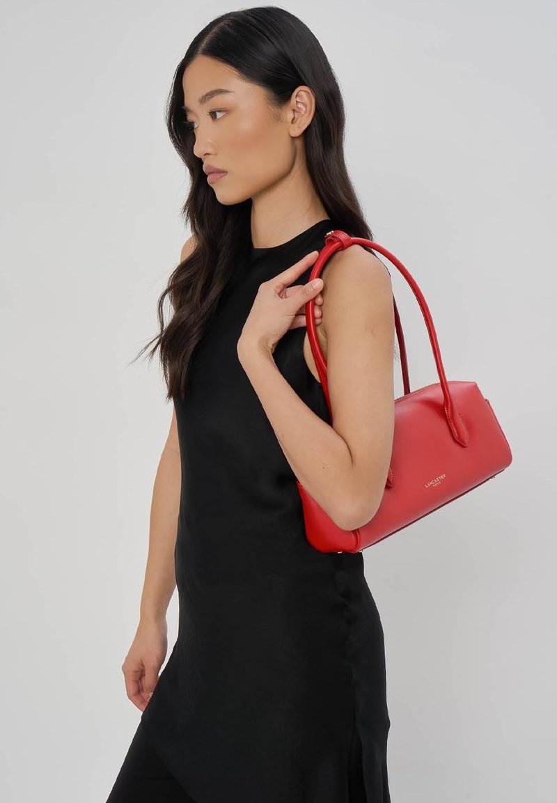 Femme en robe noire sans manches tenant un sac à main rouge Lancaster sur son épaule devant un fond uni.
