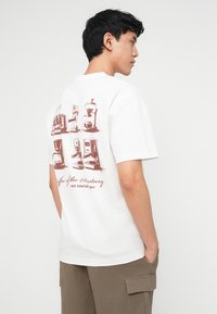 Νεαρός άνδρας που φοράει λευκό φανελάκι με καφέ εικόνες από παλαιές μηχανές καφέ και το κείμενο «Καφές του 21ου αιώνα» στην πλάτη.