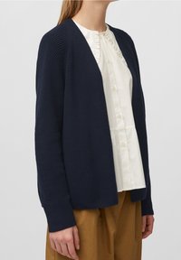 Granatowy sweter z dzianiny o teksturowanym wzorze, z otwartym przodem, ściągaczami na rękawach i dopasowanym kroju, w połączeniu z białą bluzką z falbanami.