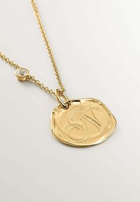 Collar de medalla dorada con un disco irregular y texturizado que presenta una letra "N" en cursiva y un pequeño acento de cristal transparente en la cadena.
