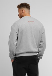 Sweatshirt gris à col rond, avec des poignets et un ourlet côtelés, arborant "UPScale Studios" imprimé en rouge au dos. Design sobre.