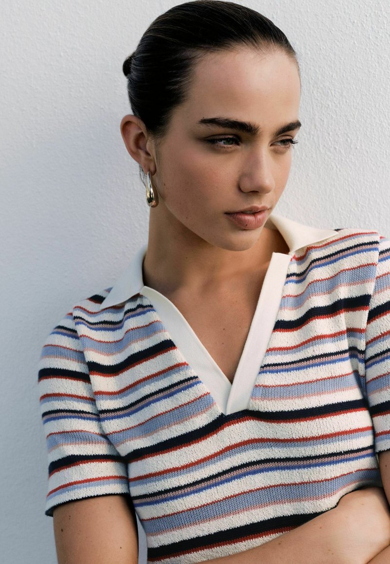 Ung kvinde med mørkt hår trukket tilbage, iført guldringearreringe og en stribet strikket kortærmet top, kigger til siden mod en hvid væg.