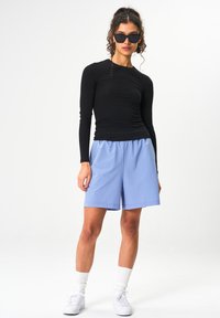 Schwarzes Langarmshirt kombiniert mit hellblauen Shorts, die über einen elastischen Bund verfügen. Das Modell trägt schwarze Sonnenbrillen und weiße Sneakers mit kurzen Socken.