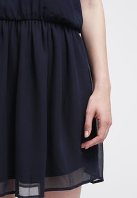 Mörkblå klänning i genomskinligt tyg med rynkad midja och flödande kjol. Sömmen är något genomskinlig, vilket visar på lager av textur. Hand vilande på kjolen.