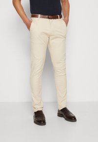 Chinos beige claro con un corte ajustado, que cuentan con bolsillos laterales y un cinturón de piel texturizada marrón. Unos mocasines marrones oscuros completan el conjunto.