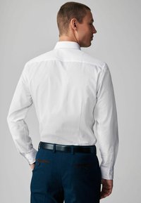 Weißes Hemd mit langen Ärmeln, Kragen und glatter Textur, kombiniert mit dunkelblauen Hosen mit braunen Taschenakzenten.