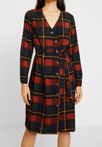 Pitkähihainen ruudullinen mekko, jossa on V-päänti, punaisia, vihreitä ja laivan sinisiä neliöitä, vyötäröllä vyö ja nappilista etuosassa.