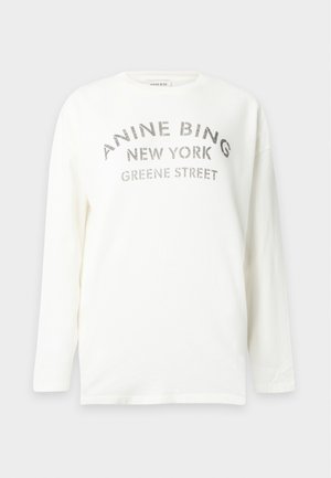Femme portant une chemise blanche à manches longues avec texte et une mini-jupe marron à carreaux, debout avec des ballerines noires sur un fond uni.