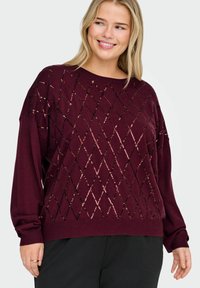 Burgundowy sweter o luźnym kroju, z diamentowymi wzorami w czarnych i różowych cekinach, długimi rękawami oraz prążkowym dekoltem i dolnym wykończeniem.