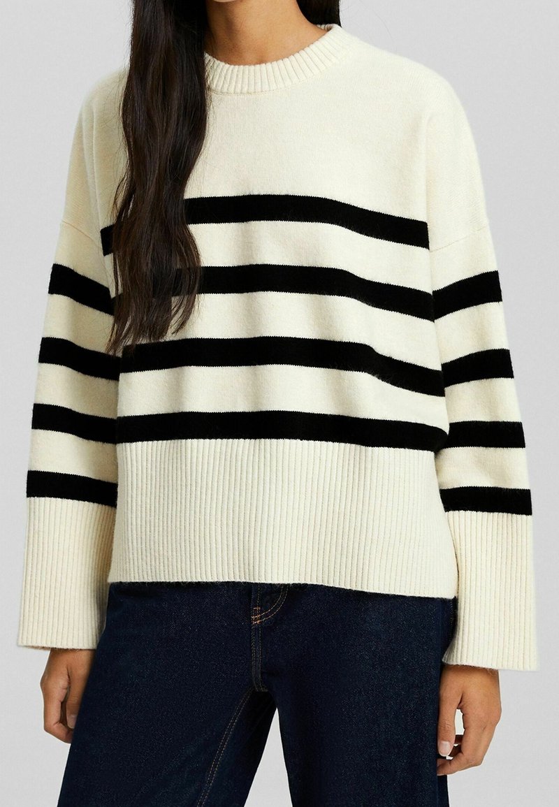 Donna che indossa un maglione lavorato a maglia crema con strisce orizzontali nere, abbinato a jeans blu scuro, in piedi davanti a uno sfondo neutro.