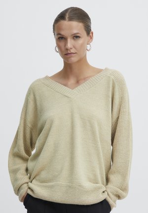 Beige, löst sittande tröja med V-ringning och texturerad stickning, ribbad mudd och fåll. Modellen bär diskreta hoopsmycken, bakgrunden är helt vit.
