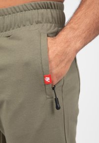 Olijfgroene broek met een elastische tailleband, voorzien van een zijzak met een rood label en een trekkoordverstelling.