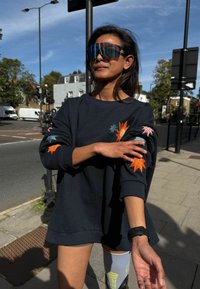 Czarna bluza z kolorowymi haftowanymi motywami słońca i gwiazd na rękawach. Model nosi odbijające światło, duże okulary przeciwsłoneczne oraz wzorzyste, sięgające do kolan skarpetki.