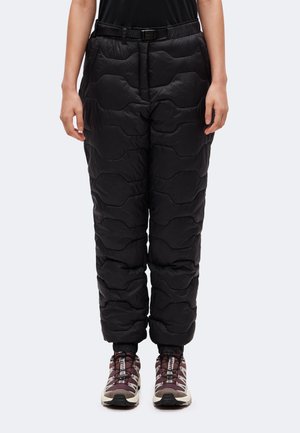 Човек, носещ черен шалтерен панталон с еластични маншети и кафяво-бели туристически обувки, стоящ срещу обикновен фон.