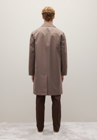 Cappotto lungo marrone con taglio dritto, dotato di uno spacco sulla schiena. Abbinato a pantaloni marroni e sneaker bianche. Design semplice e minimalista.