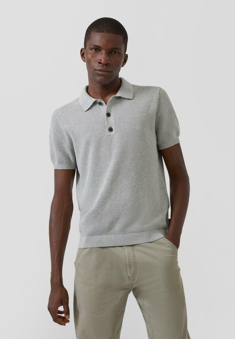 Joven de pie con una mano en el bolsillo, vistiendo un polo de manga corta texturizado color gris claro y pantalones beige, sobre un fondo liso.