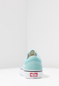 Chaussures en toile bleu clair à enfiler, avec des accents blancs et une semelle en caoutchouc noire. Présentent une étiquette de logo rouge sur le talon. Texture lisse.