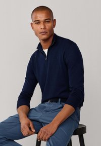 Granatowy sweter na zamek wykonany z miękkiego materiału, z długimi rękawami i wysokim kołnierzem. Nosi jasnoniebieskie spodnie, siedząc na czarnym stołku.