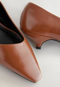 Braune Kunstleder-Pumps mit spitzer Zehenform, niedrigem Absatz und glatter Textur, ausgestattet mit einem schwarzen Innenfutter und minimalistischem Design.