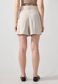 Beige shorts tinggi dengan saku samping, tekstur halus, dan potongan longgar, dipadukan dengan sepatu datar hitam.
