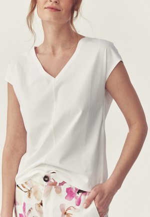 Woman wearing a white V-neck short-sleeve blouse and white pants with pink and brown floral patterns, hand in pocket, on neutral background.