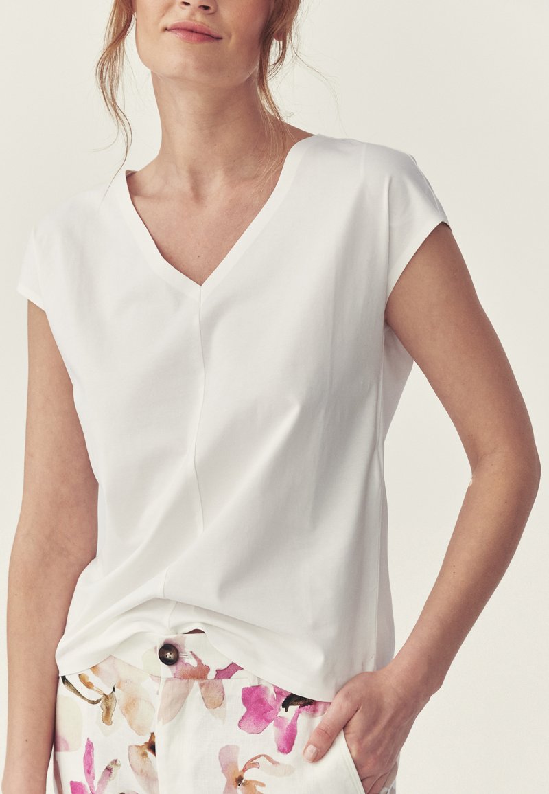 Femme portant un chemisier blanc à manches courtes et col en V et un pantalon blanc avec des motifs floraux roses et marron, main dans la poche, sur un fond neutre.