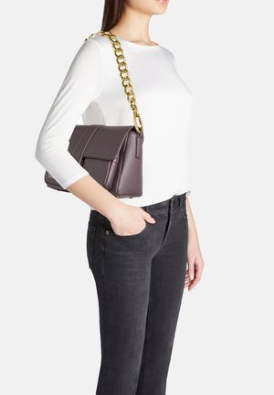 Borsa a tracolla in pelle viola con catena dorata, forma rettangolare e chiusura a pattina. Mostrata su una persona che indossa una maglietta bianca e jeans scuri.