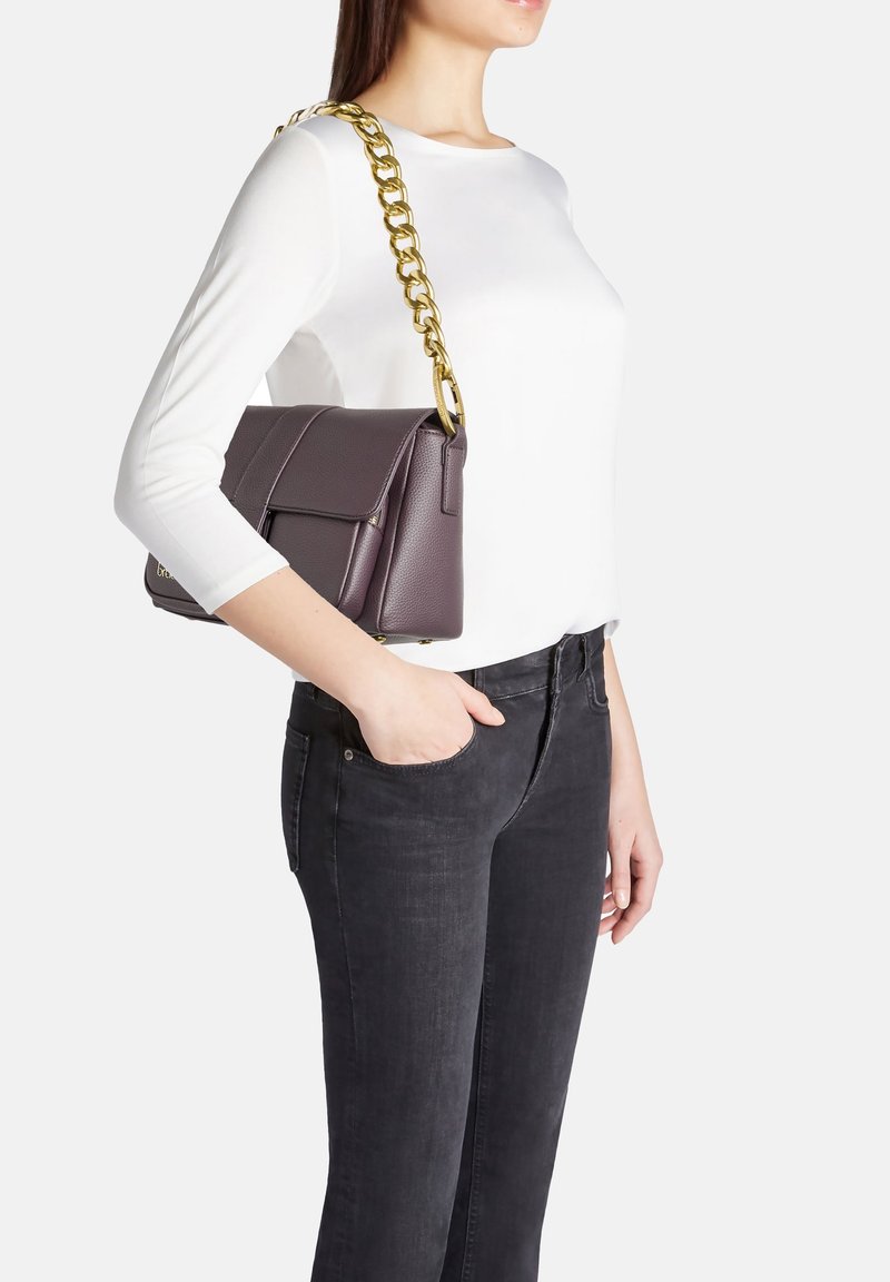 Borsa a tracolla in pelle viola con catena dorata, forma rettangolare e chiusura a pattina. Mostrata su una persona che indossa una maglietta bianca e jeans scuri.