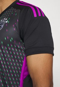 Gros plan d'un maillot de sport noir avec des motifs losanges violets et verts, des rayures violettes sur l'épaule et un logo partiel d'équipe sportive.