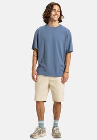 Ung mand med skulderlangt krøllet hår, iført en blå oversized t-shirt, beige shorts, stribede sokker og beige sneakers, smilende og kiggende mod højre.