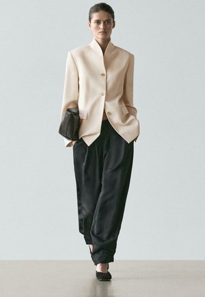 Vrouw loopt, gekleed in een beige blazer met knopen, losse zwarte broek, zwarte schoenen, en draagt een zwarte clutch onder haar arm.