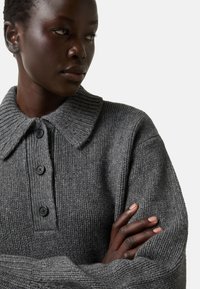 Donkerhuidige persoon met kort haar kijkt opzij, draagt een grijze gebreide trui met knopen en een brede kraag, met gekruiste armen.