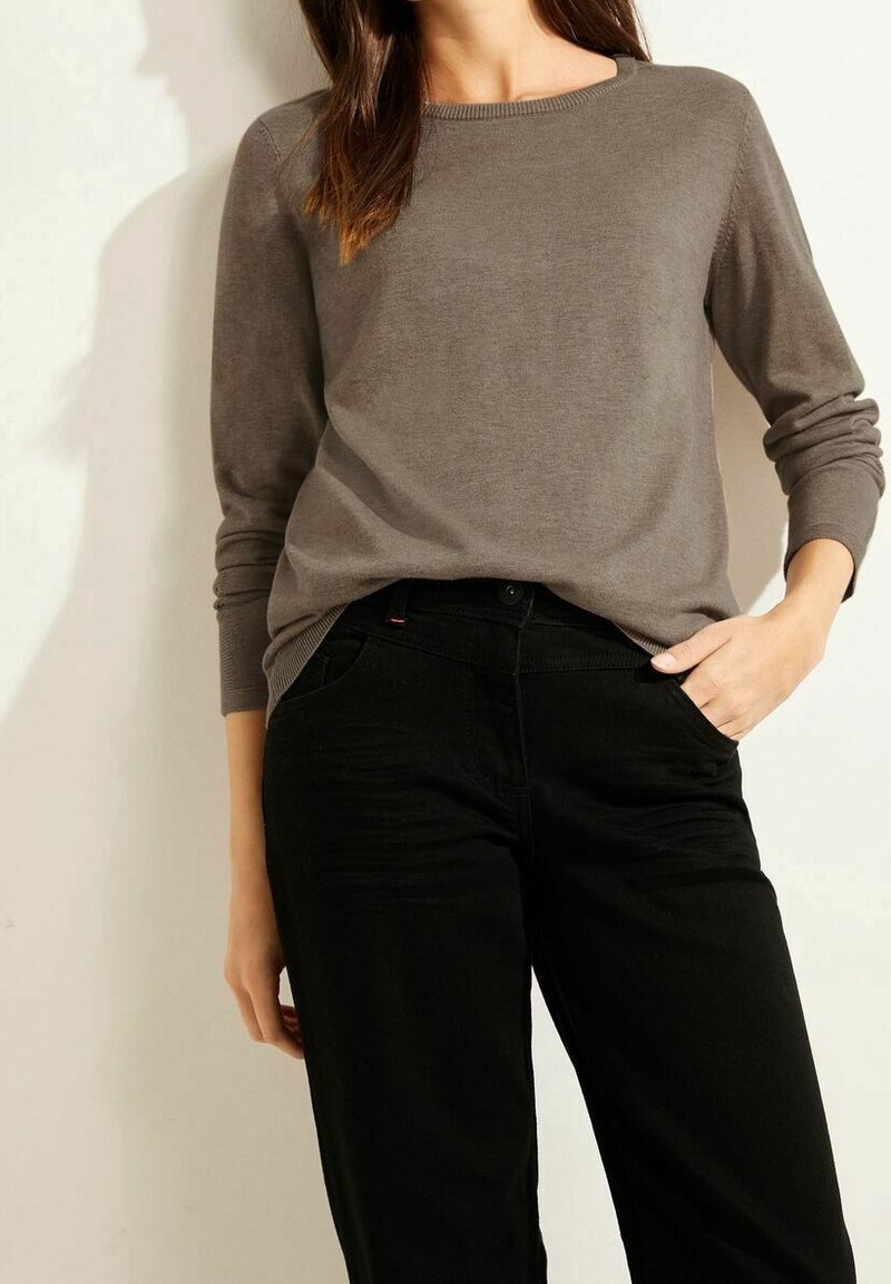 Femme portant un pull marron à manches longues rentré dans un pantalon noir taille haute, une main dans la poche, sur un fond clair.
