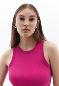 Åtsittande ärmlös linne i ljusrosa ribbat tyg, med rund halsringning och rena sömmar. Silverfärgade cirkulära örhängen framhäver looken.