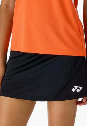 Gros plan d'une personne portant un haut orange et une jupe de sport noire avec un logo blanc Yonex en bas à droite.