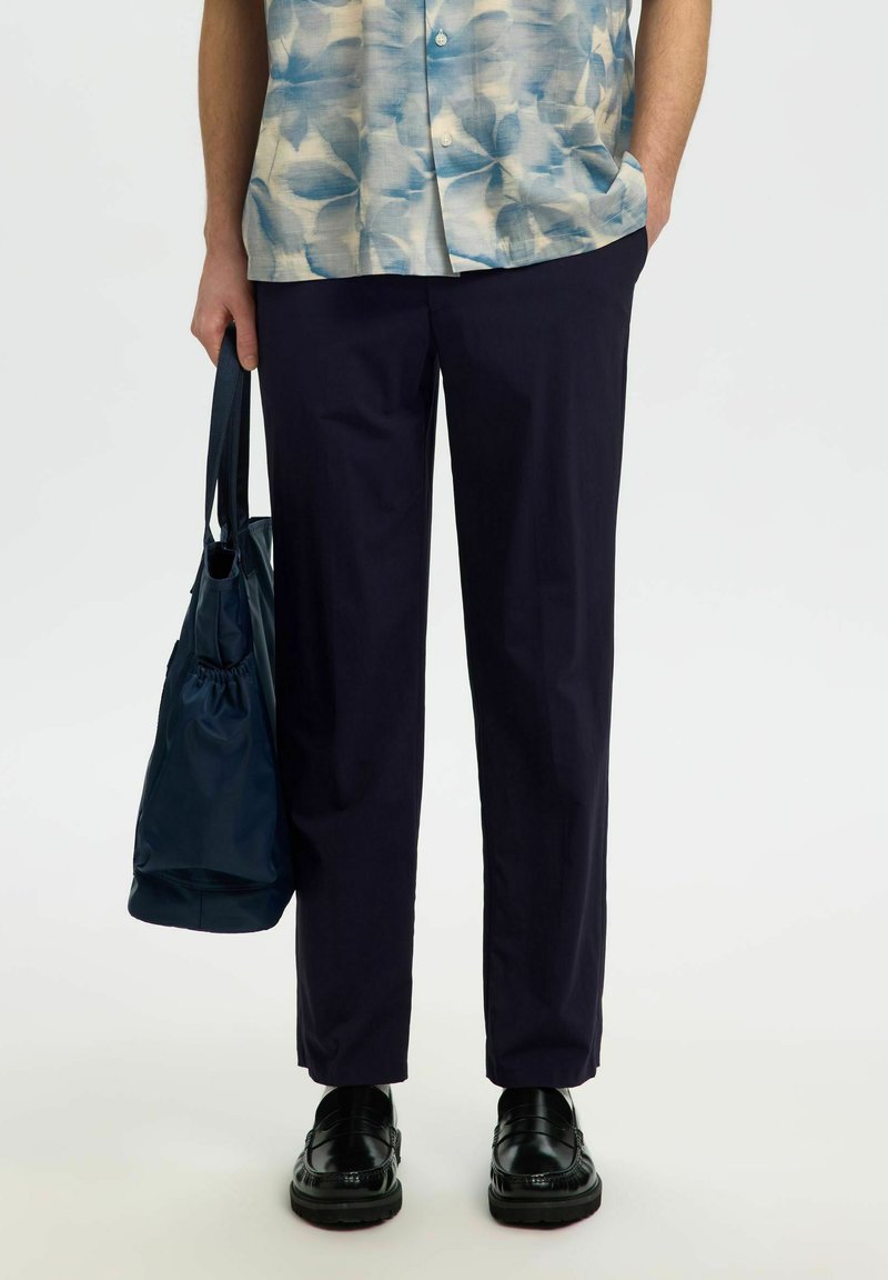 Homme portant un pantalon bleu marine, des mocassins noirs, une chemise à manches courtes bleue à motifs, tenant un sac fourre-tout bleu marine dans la main gauche.