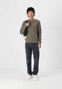 Olivengrønn strikkegenser, mørke denimjeans og hvite sneakers. Modellen bærer en svart veske med skulderrem. Enkelt design uten synlige logoer.