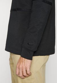 Černá mikina Nike s texturovanou tkaninou, která má výrazné logo na rukávu a tmavé akcenty. V kombinaci s béžovými kalhotami.