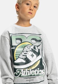 Enfant aux cheveux blonds portant un sweat gris arborant un grand motif d'une basket noir et blanc et le texte "Athletics".