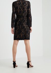 Kvinna stående med ryggen mot, iklädd en knälång svart spetsklänning med långa ärmar och svarta klacksandaler.