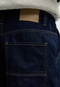 Granatowe dżinsy mają gładką teksturę, żółte przeszycia i skórzaną naszywkę w talii z wytłoczonymi detalami.