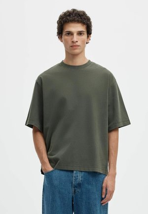 Giovane uomo con capelli ricci che indossa una t-shirt verde scuro oversize e jeans blu, in piedi con una mano in tasca.